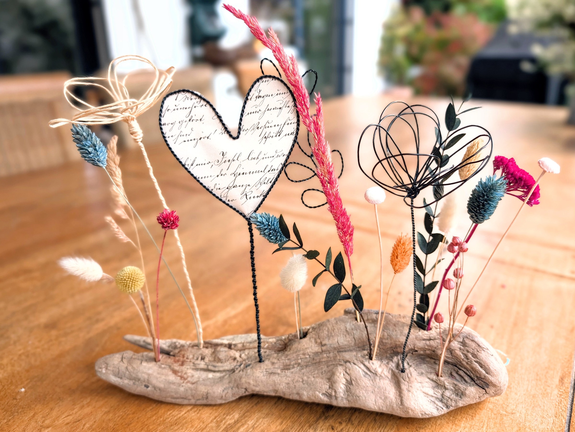 Drahtherz mit Papiereinlage und Trockenblumen auf Treibholz, auf braunem Holztisch